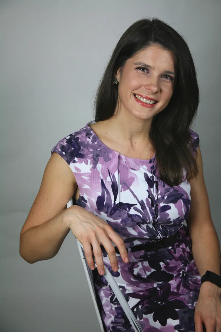 Person in a floral dress sitting on a chair, smiling at the camera against a plain background.