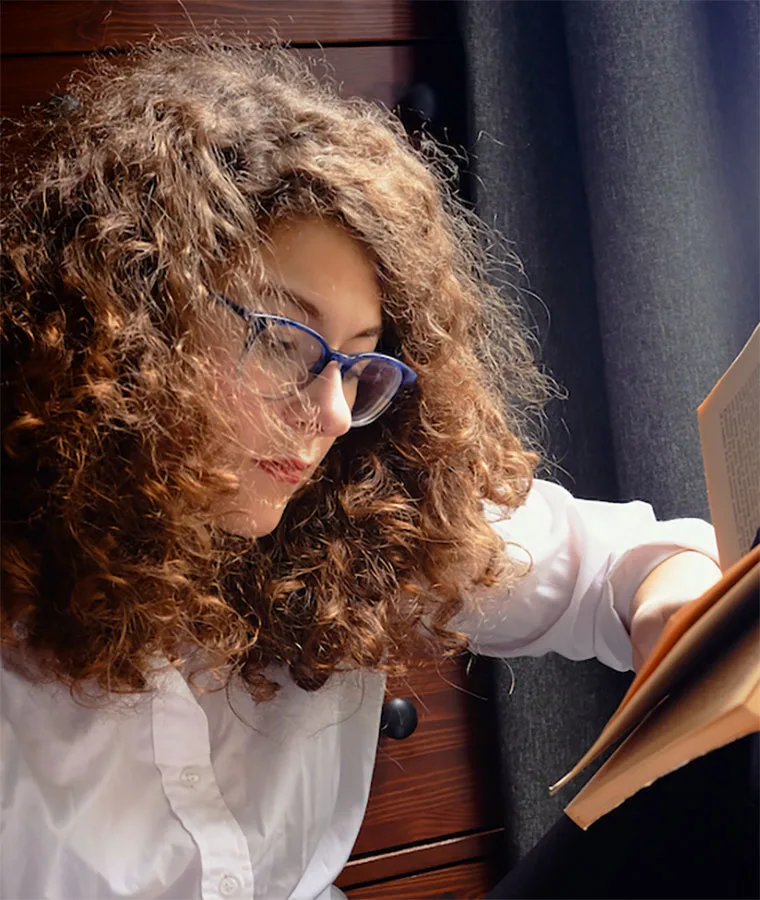 Curly-haired person wearing glasses and a white shirt, sitting indoors by a dark curtain, reading a book.
