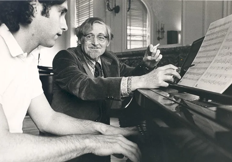 Two individuals at a piano, one playing while the other gestures at sheet music, engaging in a musical discussion.