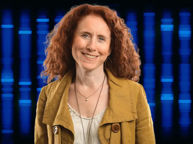 Person with curly red hair wearing a mustard jacket, smiling in front of a blue vertical light pattern background.