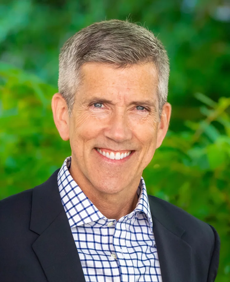 Smiling person with short gray hair, wearing a checkered shirt and dark blazer, against a blurred green background.
