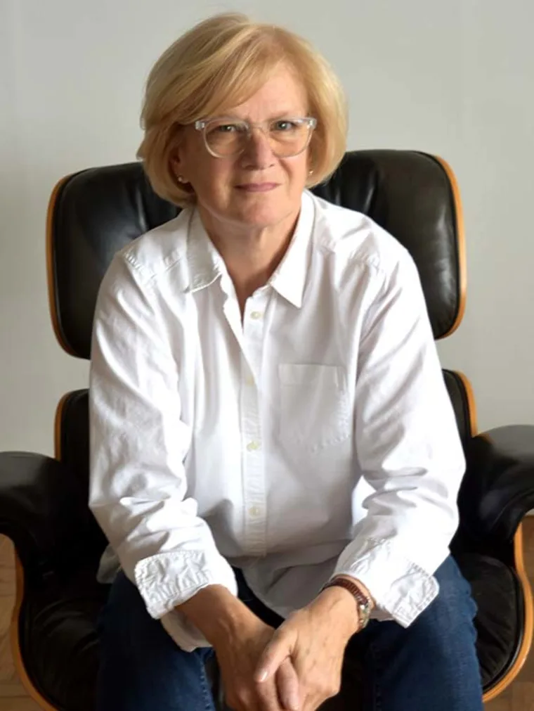 Person with short blonde hair and glasses, wearing a white shirt, sits on a black leather chair, hands clasped, looking forward.