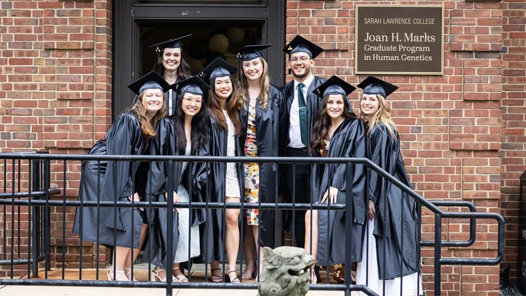 Group of graduates in caps and gowns pose on steps in front of a brick building with a sign reading "Joan H. Marks Graduate Program in Human Genetics."