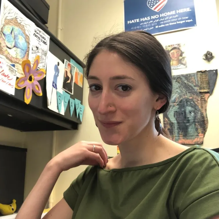 Person sitting at a desk with art and posters on the wall. A sign reads "Hate has no home here" in multiple languages.