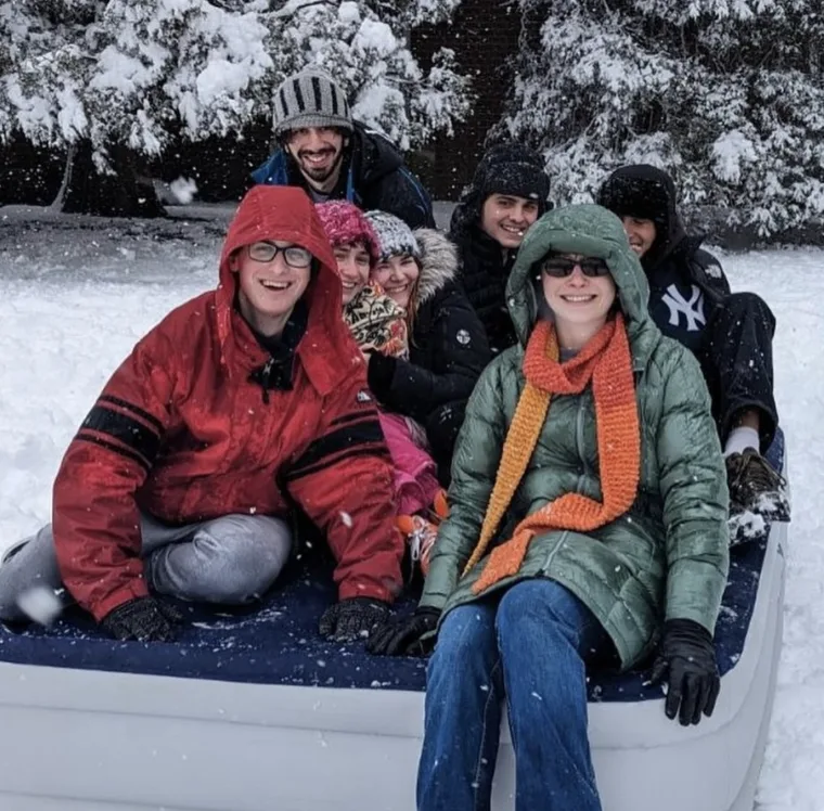 Group of people in winter clothing sitting on a snow-covered inflatable sled, smiling with trees in the background.