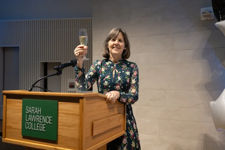 Person in floral outfit holds a champagne glass, standing at a podium labeled "Sarah Lawrence College."