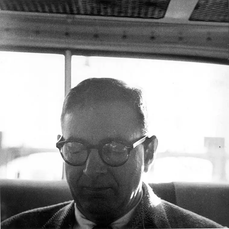 Person with glasses, seated inside a vehicle, looking down. Bright light from the window behind.