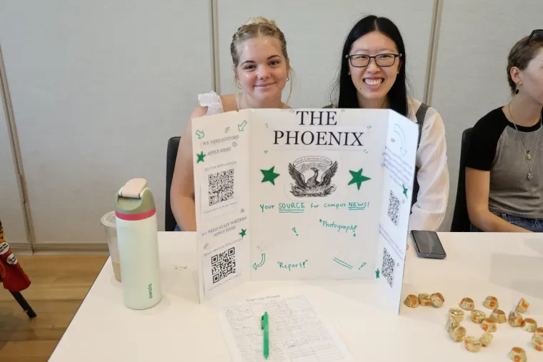 Two people sit behind a table with a display board titled "The Phoenix," featuring information, QR codes, a signup sheet, and small snacks on the side.
