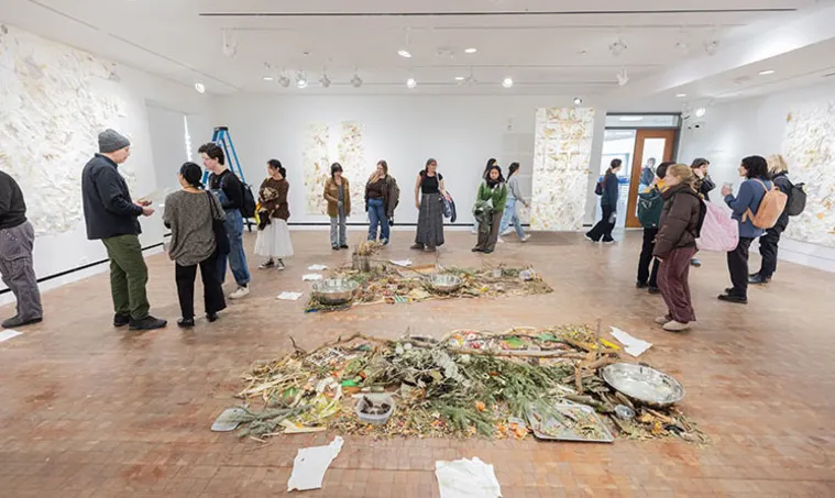 People in a gallery observe art installations on the floor and walls, with scattered foliage and objects, and bright lighting overhead.
