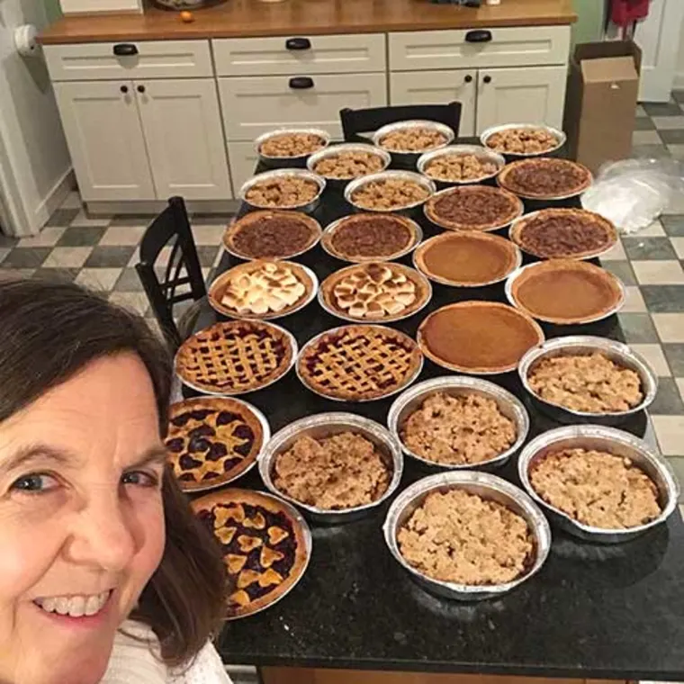 Person smiling in a kitchen with a table full of various pies, including lattice-topped and crumb-topped varieties.