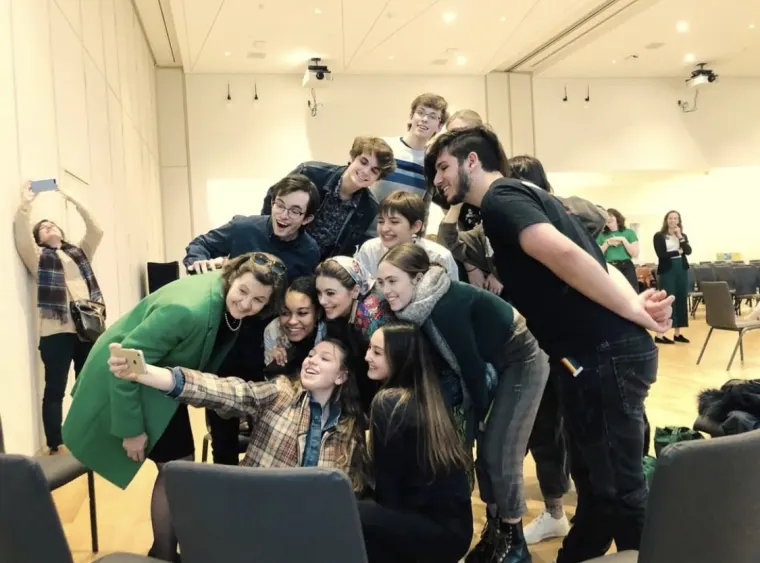 A group of people gathers closely for a selfie in a bright room, with one person in the background stretching and taking a photo.