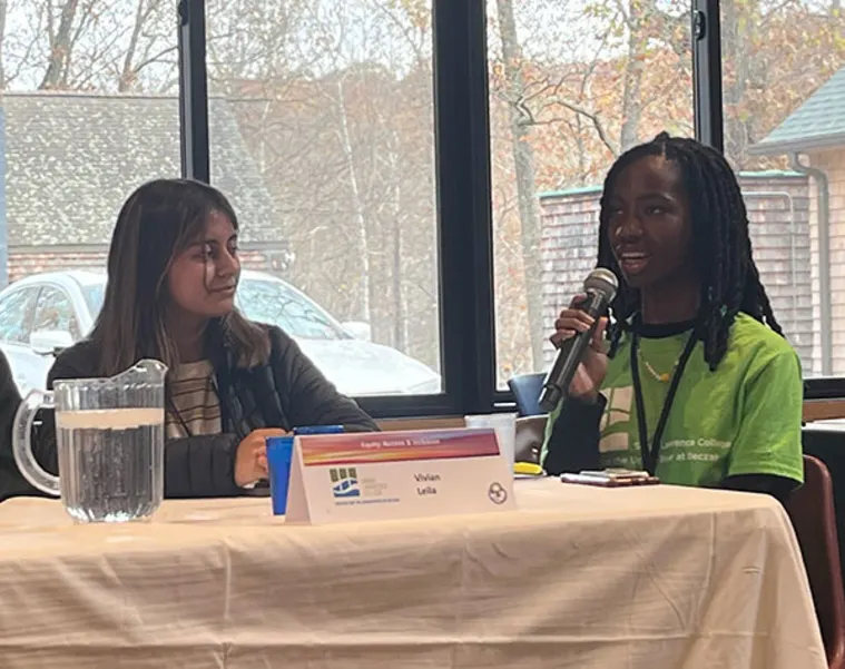 Two people sit at a table with a microphone and name cards, engaged in a discussion. A window with a view outside is in the background.