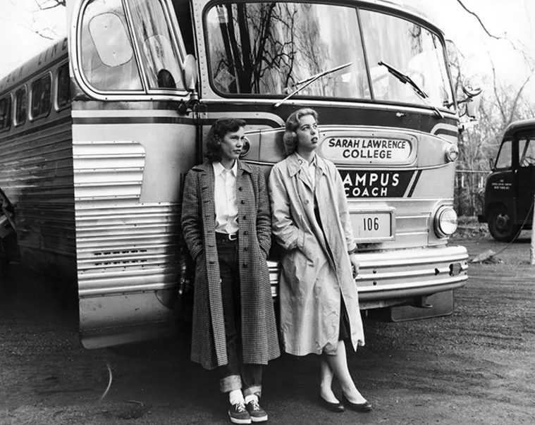 Two people in long coats stand in front of a vintage bus labeled "Sarah Lawrence College" on a campus road.