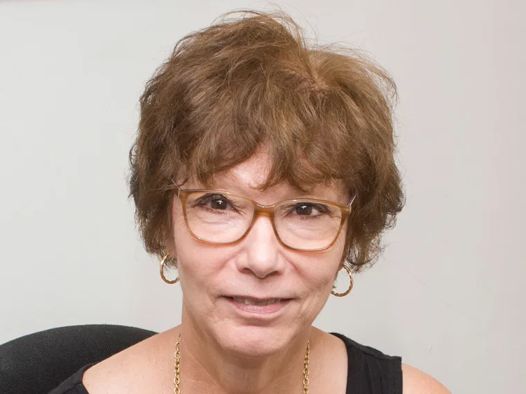 Person with short, curly hair wearing glasses, gold earrings, and a necklace, smiling slightly, with a neutral background.