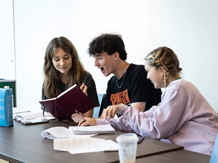 Three people sit at a table, engaged in discussion. One reads from a book, another laughs, and the third takes notes. Papers and a drink are on the table.