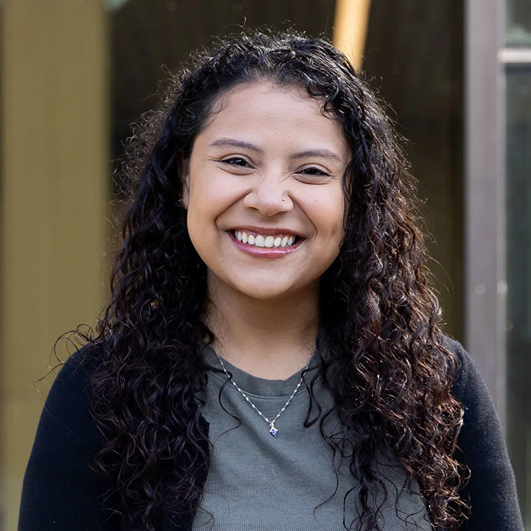 Person smiling with long, curly hair, wearing a necklace and a dark sweater over a green top, standing outdoors.