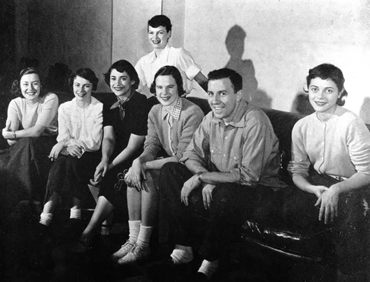 Seven people sitting and smiling, with one standing in the back. They are dressed in mid-20th century casual clothing.