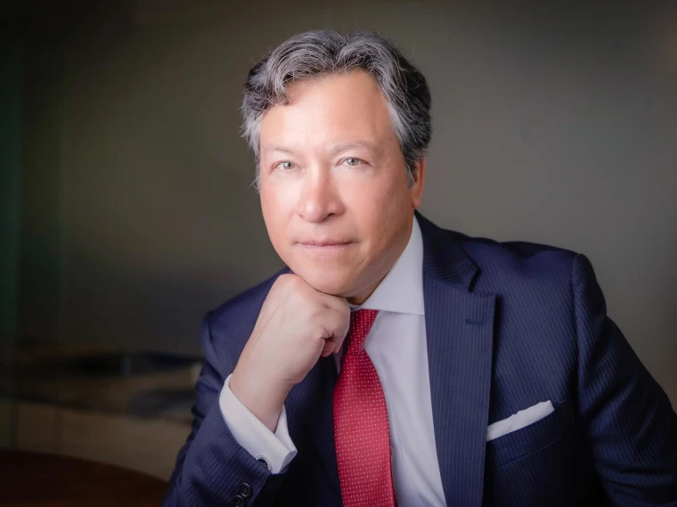 Person in a suit with a red tie, resting chin on hand, gazes forward. Background is softly blurred, creating a professional portrait setting.
