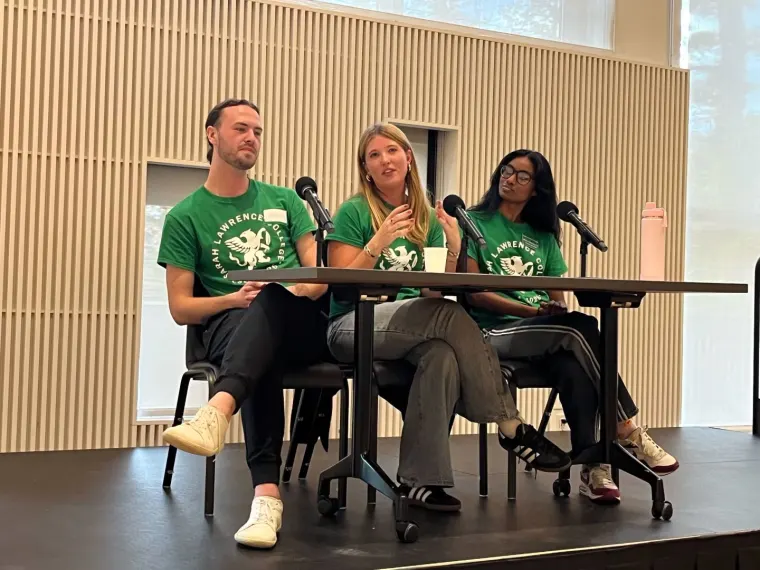 Three people sitting at a table wearing matching green shirts with microphones in front of them, engaged in a discussion.