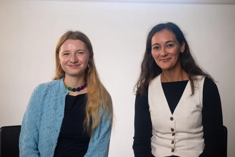 Two people are sitting and smiling at the camera against a plain background. One wears a blue sweater, the other a white vest over a black shirt.
