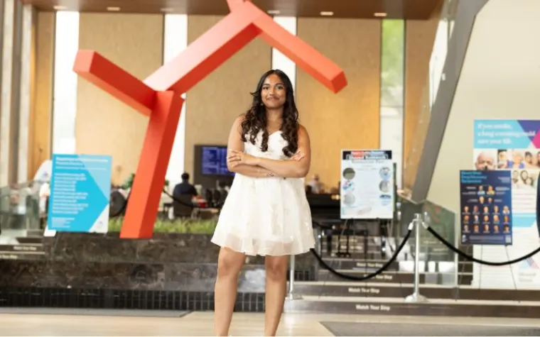 Person in white dress stands confidently indoors in front of a large red abstract sculpture, surrounded by informational posters.