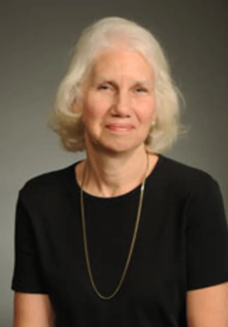 Individual with short white hair, wearing a black top and a thin gold necklace, against a plain background.