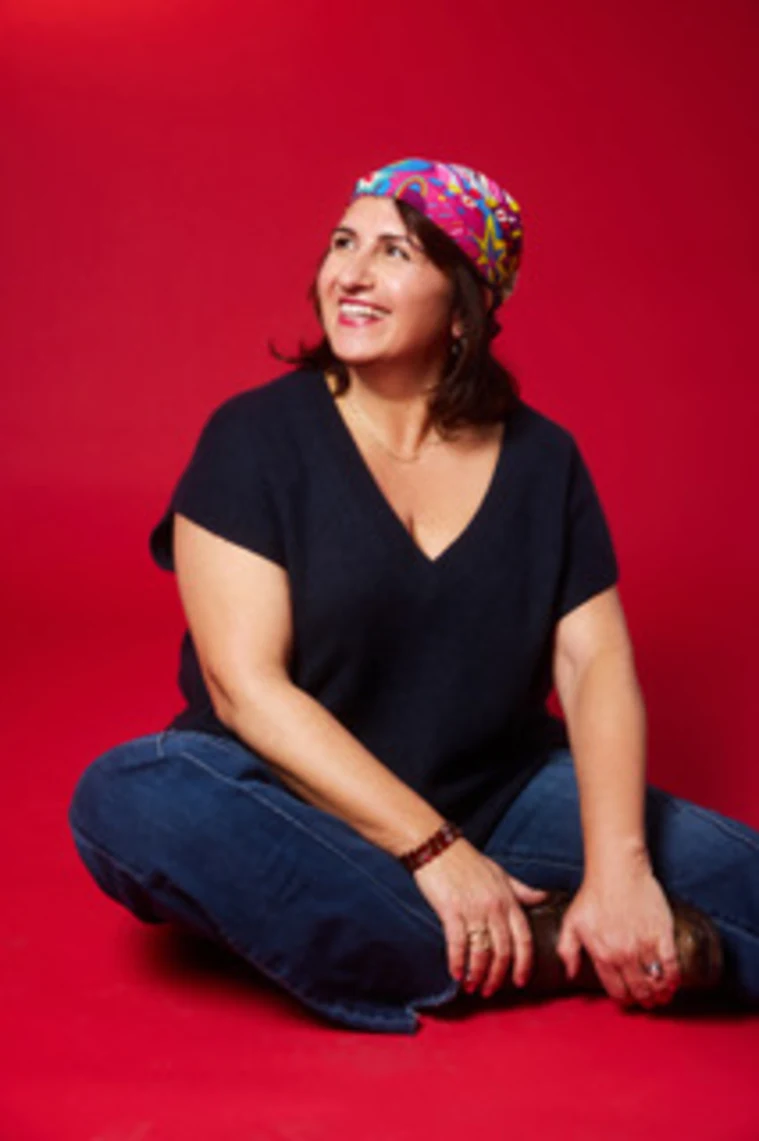 Person sitting cross-legged on a red background, wearing a colorful headscarf, navy shirt, and jeans, looking to the side and smiling.