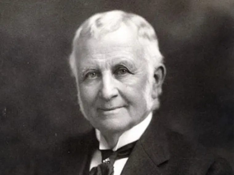 Older person with short white hair and sideburns, dressed in a dark suit and white shirt, smiling slightly in a vintage black-and-white portrait.