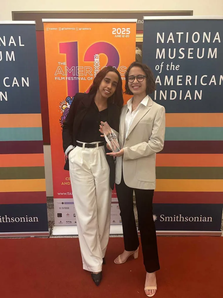 Two people stand smiling, holding an award between them in front of a colorful film festival banner at the National Museum of the American Indian.