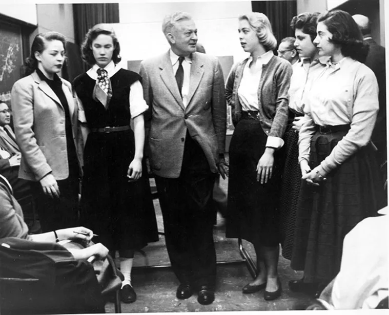 A group of six individuals gathers in a room, with one person in a suit speaking to the others who are wearing skirts and blouses.