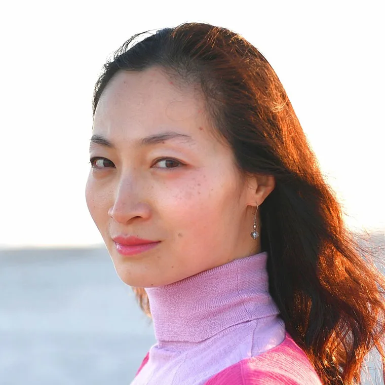 Person wearing a pink turtleneck and earrings, with long hair, standing outdoors with the sun shining on the face.