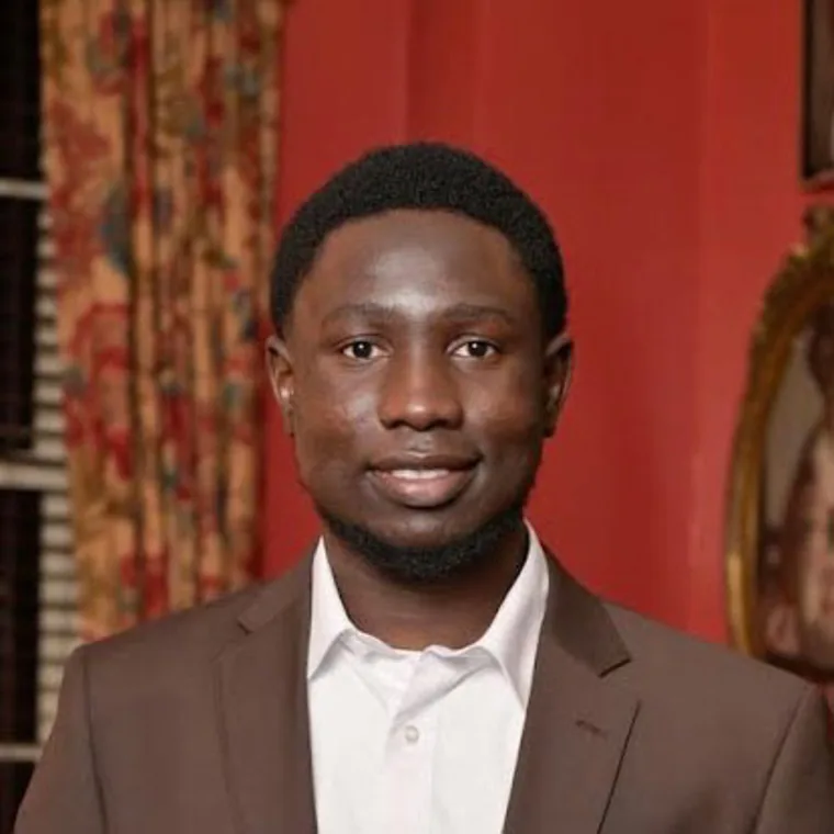 Person in a brown suit and white shirt smiling in a room with red walls and floral curtains.