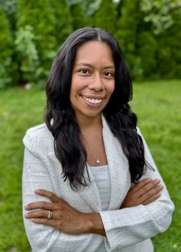 Person with long hair smiling, arms crossed, in a white jacket, standing outside with greenery in the background.