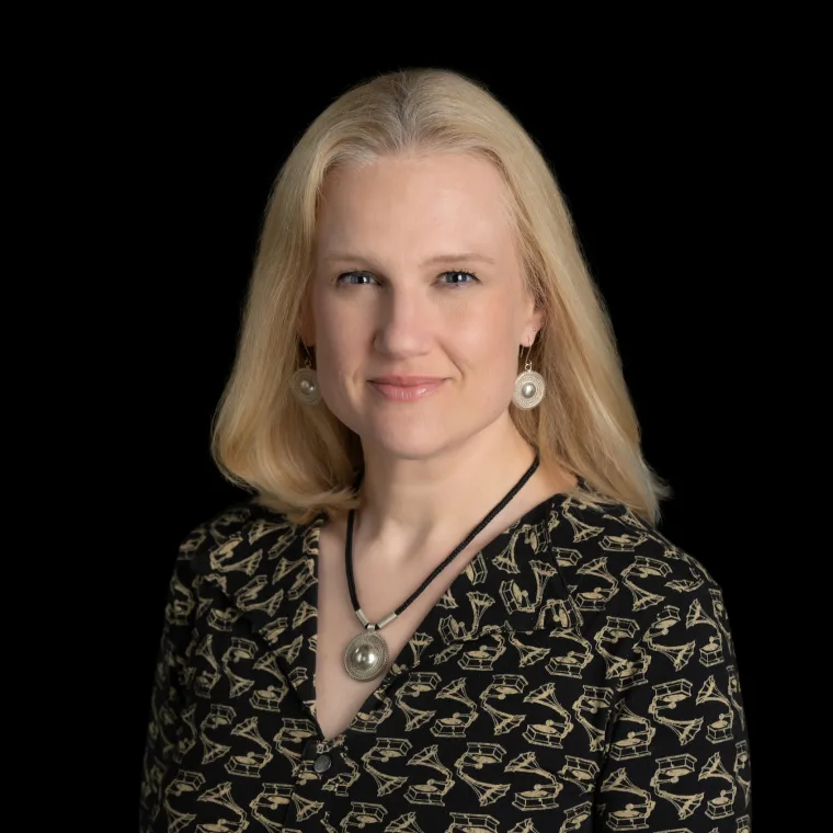 Person with light hair wearing a patterned shirt and large round earrings, smiling at the camera against a black background.
