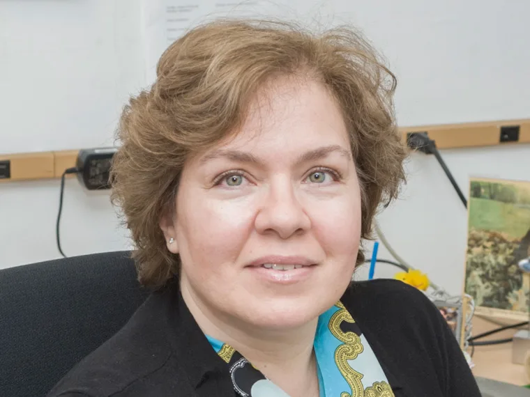 Person with short, curly hair sits in an office with a neutral expression. Background includes a wall, electrical outlets, and a framed picture.