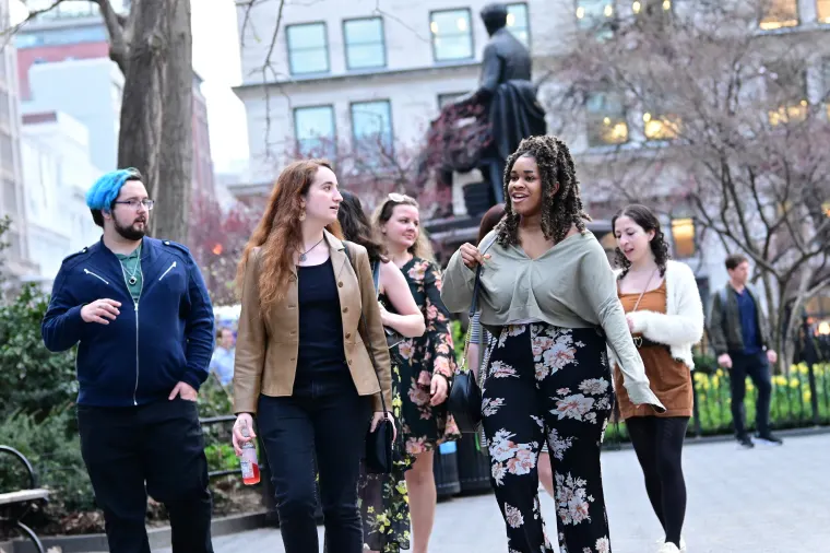 Group of people walking and chatting in a park with trees and a statue in the background, surrounded by buildings.