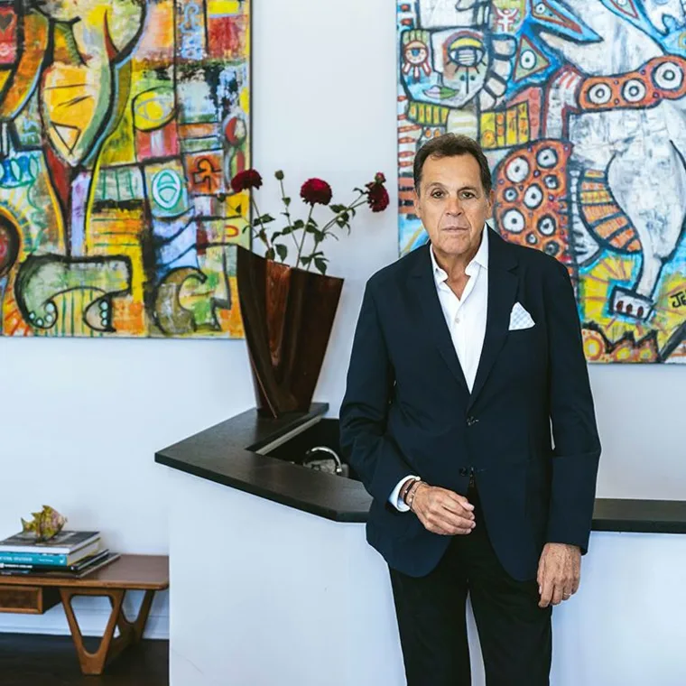 Person in a suit stands in a room with vibrant abstract paintings and a wooden vase with flowers on a counter.