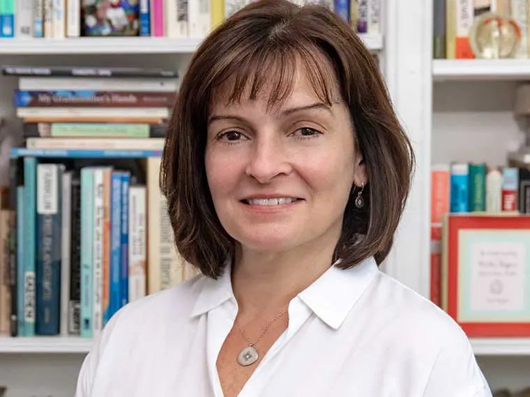 Person with short brown hair smiling in front of a bookshelf filled with various colored books, wearing a white shirt and necklace.