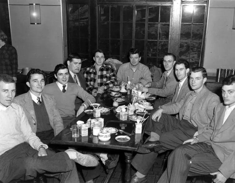 A group of individuals seated around a table with dishes, drinks, and food in a dining setting, wearing a mix of casual and formal attire.