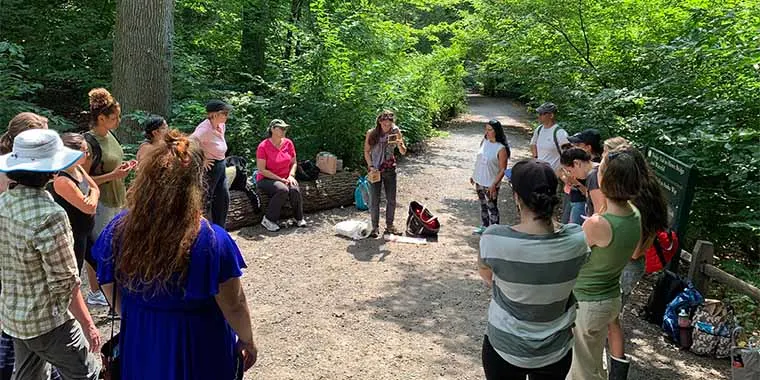 A group of people stands in a wooded area, listening to someone. Backpacks and personal items are on the ground nearby.