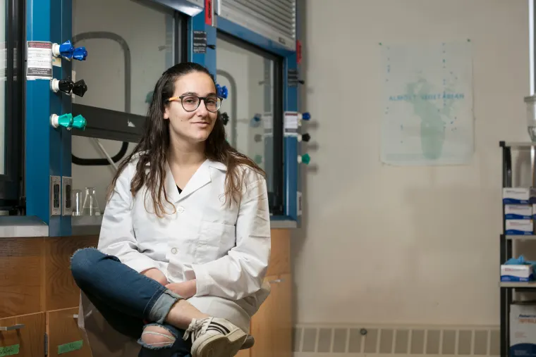 Person wearing a lab coat and glasses sits casually on a counter in a laboratory setting with equipment and a poster in the background.