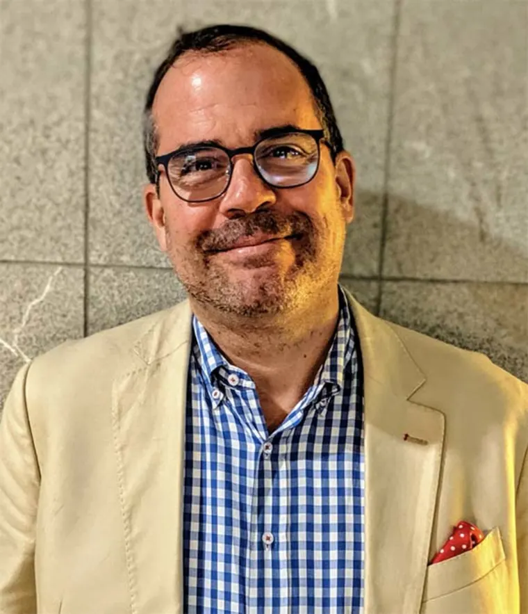 Person with glasses, wearing a beige jacket and blue checkered shirt, stands against a tiled wall, smiling slightly.