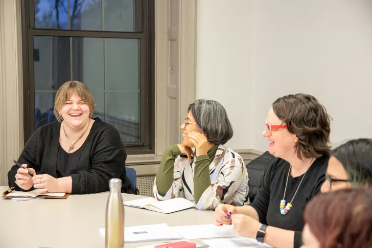 Group of people sitting around a table, one person laughing, others engaging in conversation, papers and notebooks on the table.