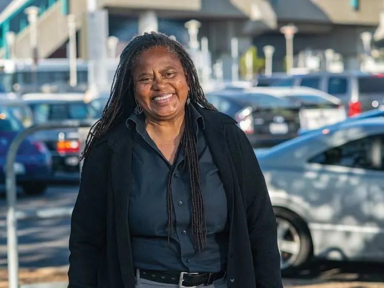 Person with long braided hair smiling outdoors, wearing a black shirt and jacket. Cars and buildings are visible in the background.