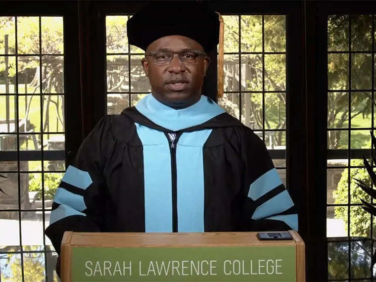 Person in academic regalia stands at a podium labeled "Sarah Lawrence College," with a window and trees in the background.