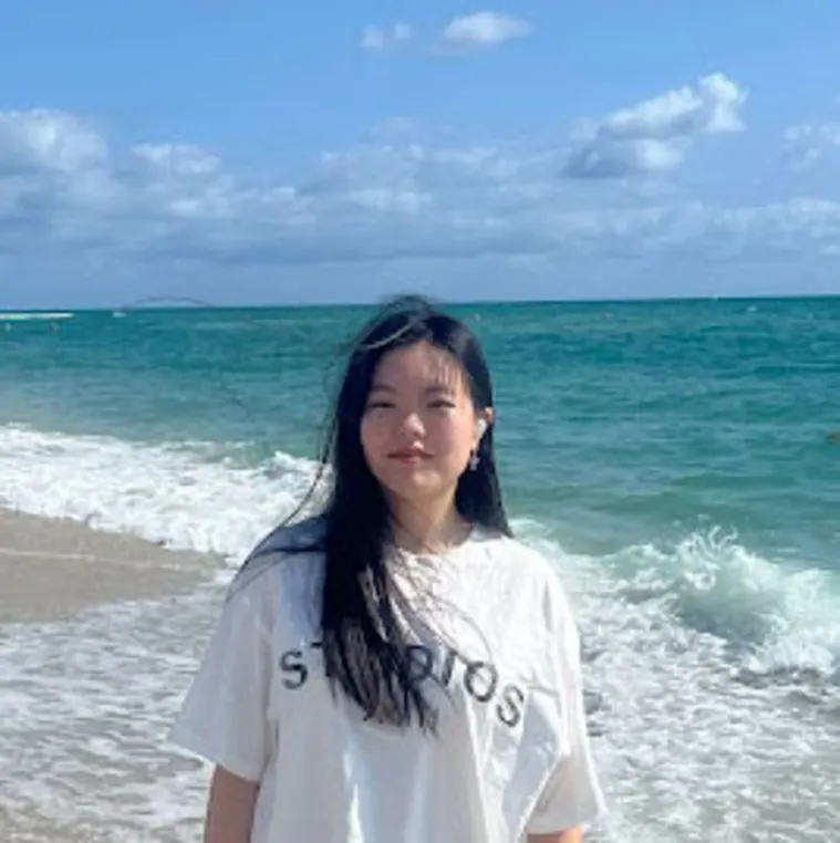 Person in a white T-shirt stands on a beach with waves and a blue sky in the background.