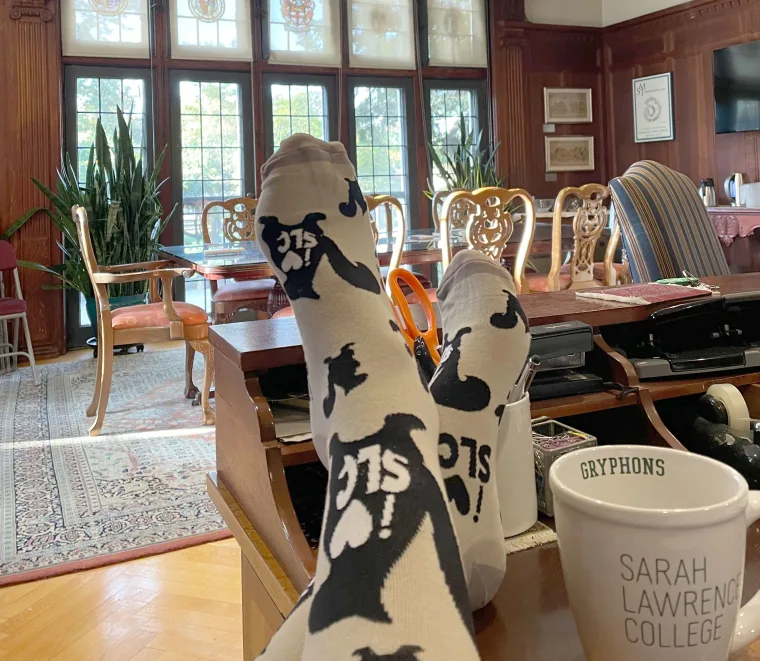 Feet in white socks with black designs rest on a desk next to a mug labeled "Gryphons Sarah Lawrence College" in a wood-paneled room.