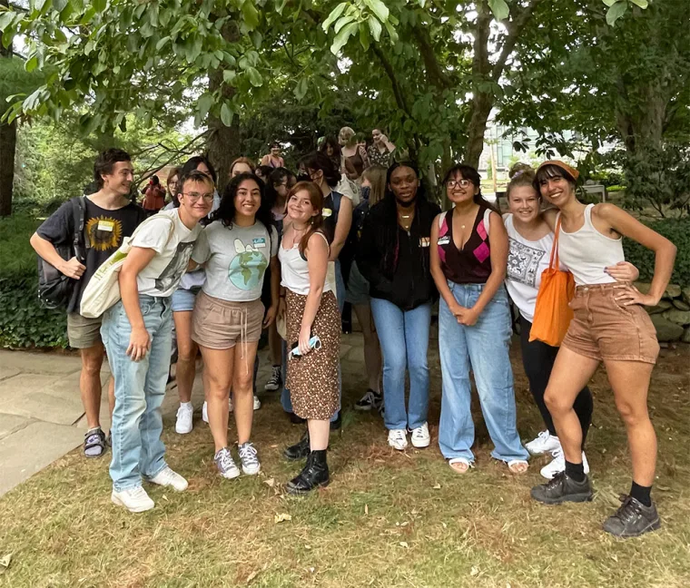 A group of people smiling and standing together under a tree in a park setting, with some carrying bags.