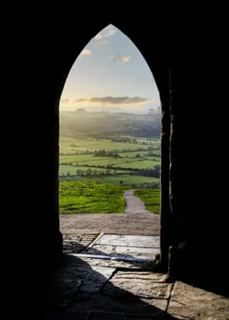View of a green landscape at sunrise seen through a stone archway, with a pathway leading into the distance.