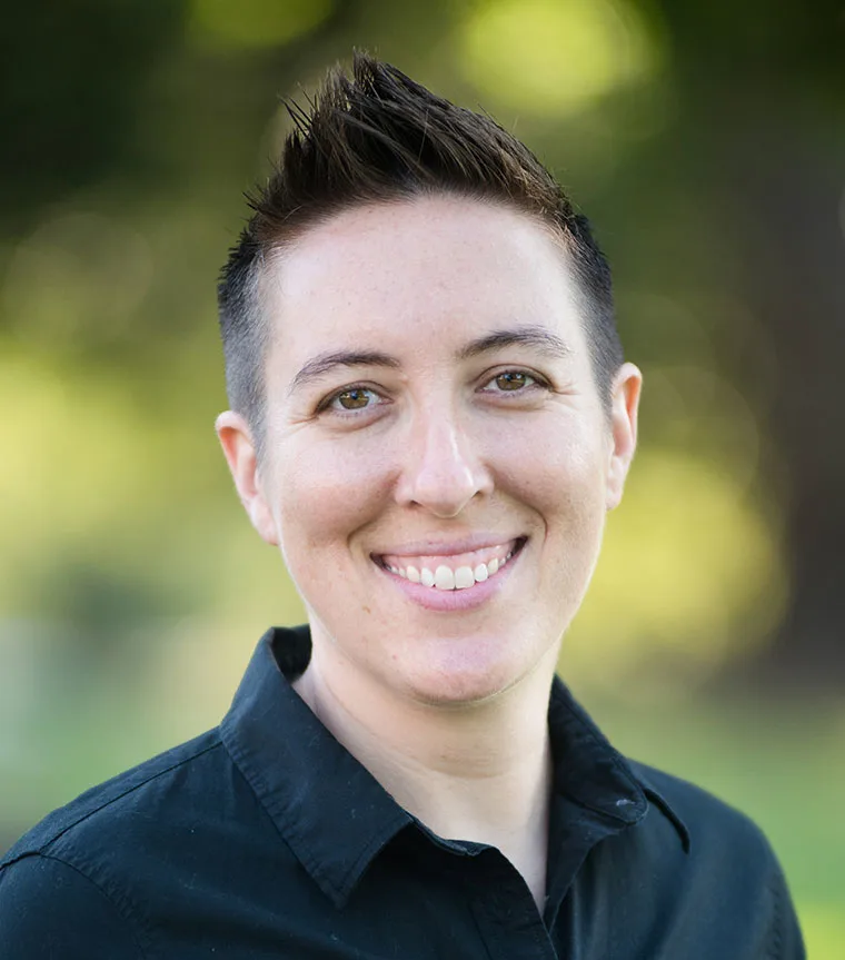 Person with short hair smiling, wearing a black collared shirt. Blurred green and yellow background outdoors.